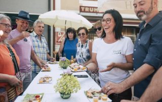 Gruppe beim Besuch des 1. Stopps am kulinarischen Spaziergang: Michaela Stangl präsentiert am Tisch mit weißem Tischtuch Bauernmarkt Brötchen und Vitamin-Shots.