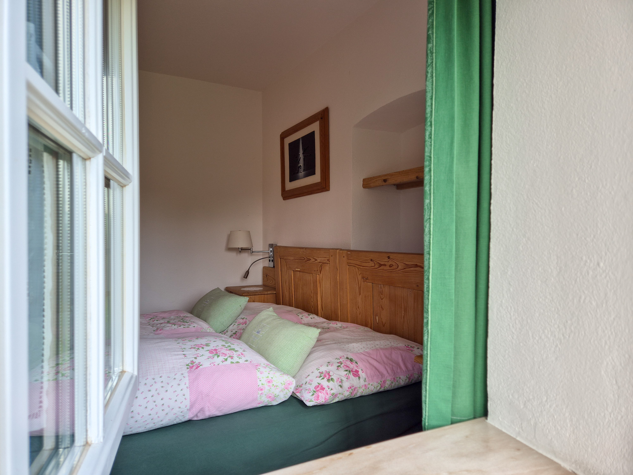 Durch ein geöffnetes Fenster fällt der Blick auf ein Doppelbett mit rosa-weißem Bezug