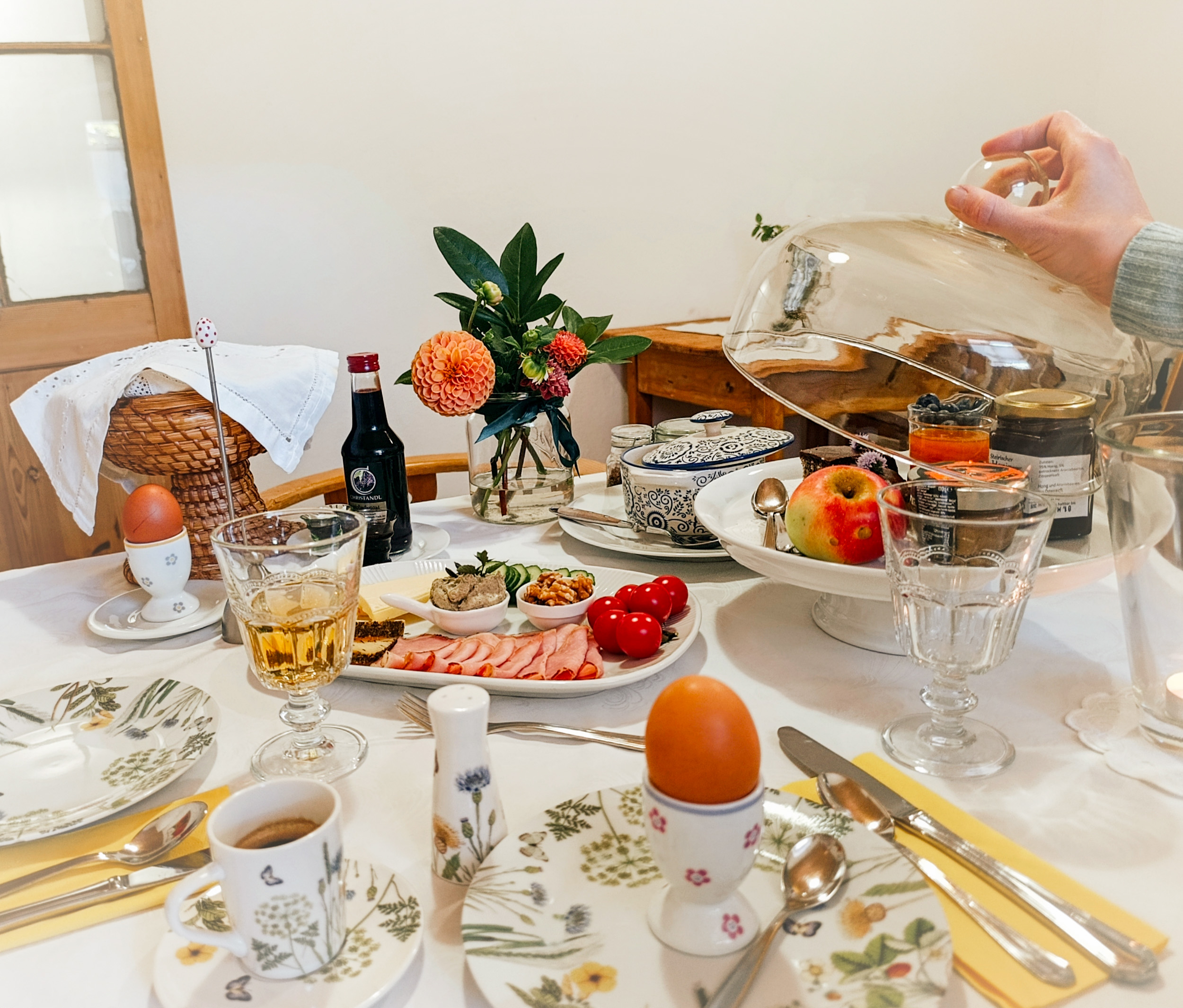 Auf einem Tisch steht ein Frühstück mit Ei, Saft, Schinken-Käse-Platte und Brotkorb. Eine Hand öffnet die Glaskuppel, unter der süße Sachen zu sehen sind.