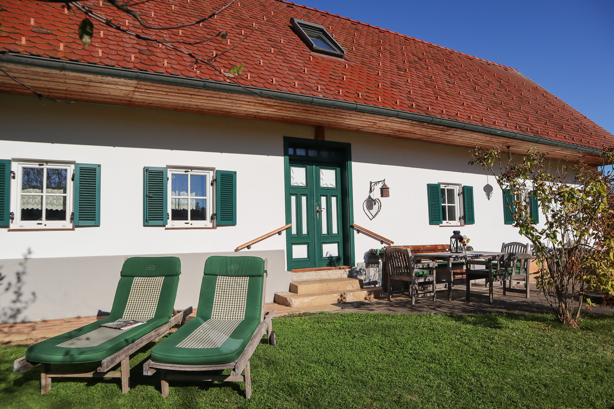 Zwei Holzliegen mit grünen Auflagen stehen im Garten eines weiß gestrichenen Landhauses mit grünen Balken, grüner Eingangstür und rotem Ziegeldach